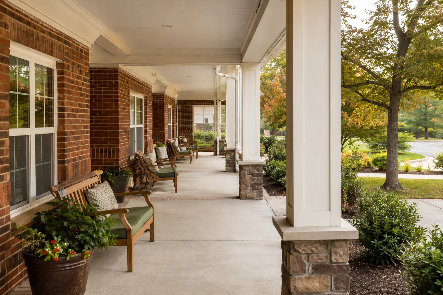 A covered veranda with benches along a brick care-community building, looking onto mature trees